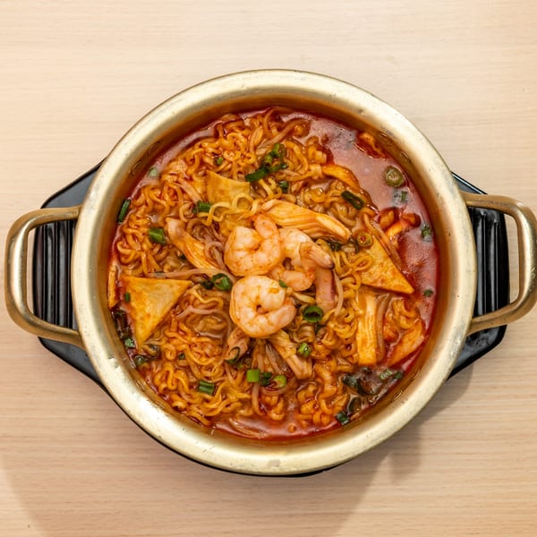 Top-down view of a golden pot filled with spicy ramen, shrimp, and vegetables on a wooden table.