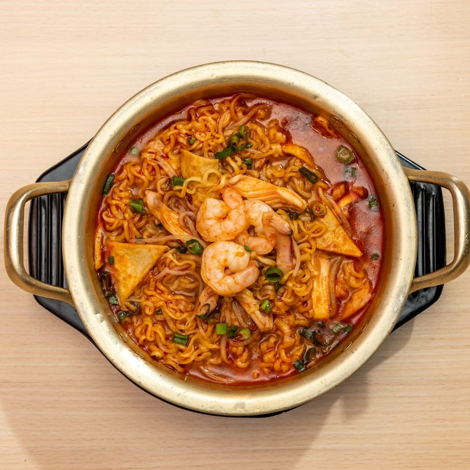 Top-down view of a golden pot filled with spicy ramen, shrimp, and vegetables on a wooden table.