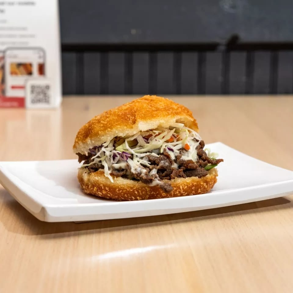 A Korean bulgogi sandwich with shredded cabbage and sauce on a crispy bun, served on a white plate.