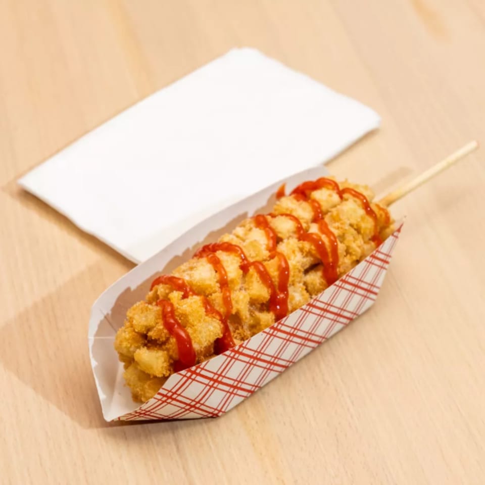 Korean corn dog with crispy potato cubes and ketchup drizzle in a paper tray on a wooden table.