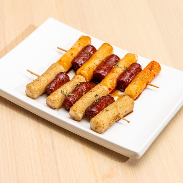 Skewers of grilled tteokbokki and sausages, drizzled with sauce and herbs, on a white plate.