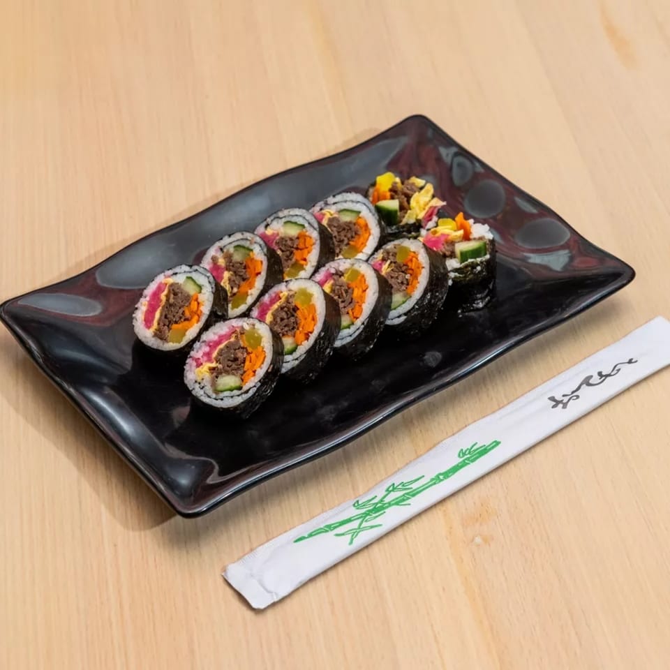 A black rectangular plate holds ten pieces of Korean gimbap with chopsticks on a wooden table.